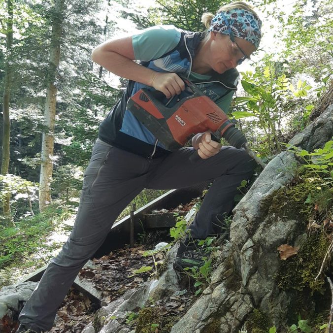 Frau im Wald bohrt mit einer Bohrmaschine in den Berg um Bohrhaken zu befestigen.